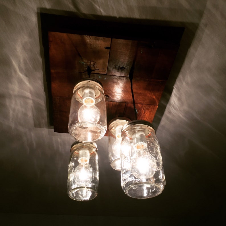 May include: A rustic wooden ceiling fixture with four glass jars used as light fixtures. The jars have clear glass and are lit with warm white light bulbs.