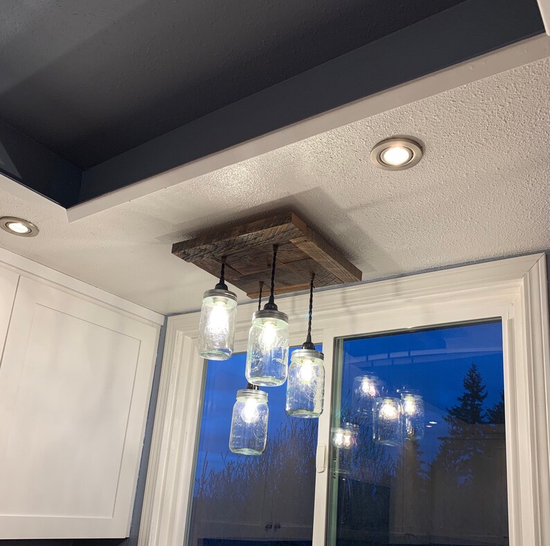 May include: A rustic wooden ceiling light fixture with five glass jars as light shades. The jars are clear glass and have a frosted finish. The fixture is hanging from the ceiling and is illuminated. The light fixture is in a room with a window in the background.