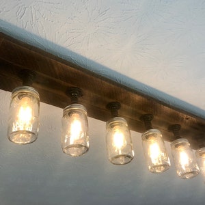 Pu&ograve; includere: Un lampadario rustico in legno per soffitto con sei barattoli di vetro trasparente e tubi di metallo a vista. I barattoli sono illuminati da lampadine a luce bianca calda.