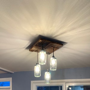 May include: A rustic wooden ceiling light fixture with four glass mason jar pendant lights. The fixture is made of reclaimed wood and has a natural finish. The mason jars are clear glass and have a simple design.