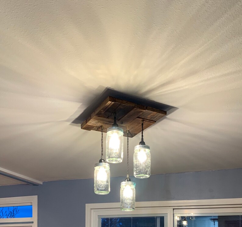 May include: A rustic wooden ceiling light fixture with four glass mason jar pendant lights. The fixture is made of reclaimed wood and has a natural finish. The mason jars are clear glass and have a simple design.
