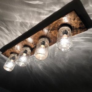 Könnte beinhalten: Eine rustikale Deckenleuchte aus Holz mit vier durchsichtigen Glas-Einmachgläsern, die jeweils eine Glühbirne enthalten. Die Gläser sind mit Metallbefestigungen am Holz befestigt.