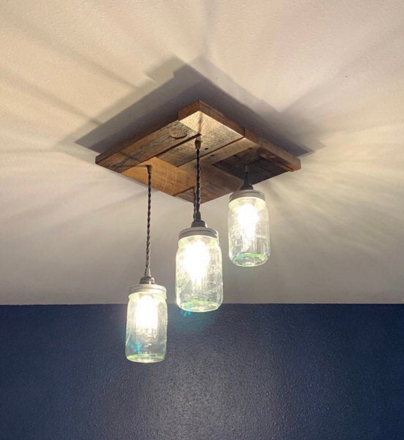 May include: A rustic ceiling light fixture made from reclaimed wood with three mason jar pendant lights. The jars are clear glass with a light bulb inside each one.