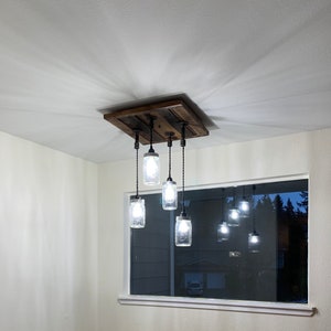 Könnte beinhalten: Eine rustikale Deckenleuchte aus Holz mit sechs Glasbehältern, die daran hängen. Die Behälter haben Glühbirnen im Inneren und sind beleuchtet. Die Leuchte ist an der Decke montiert und hat eine dunkelbraune Oberfläche.