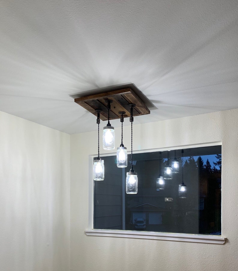 May include: A rustic wooden ceiling light fixture with six glass jars hanging from it. The jars have light bulbs inside and are lit up. The fixture is mounted to the ceiling and has a dark brown finish.