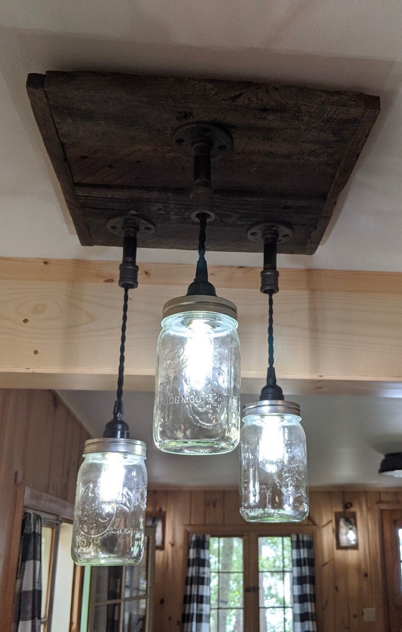 May include: A rustic ceiling light fixture made from reclaimed wood with three mason jar lights hanging from black cords.