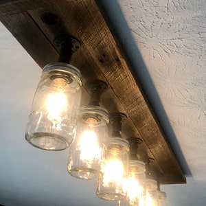 Pu&ograve; includere: Un apparecchio di illuminazione rustico in stile farmhouse con sei barattoli di vetro attaccati a una tavola di legno. I barattoli sono cablati per lampadine e sono sospesi da tubi di metallo nero.