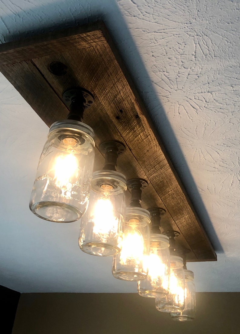 Pu&ograve; includere: Un apparecchio di illuminazione rustico in stile farmhouse con sei barattoli di vetro attaccati a una tavola di legno. I barattoli sono cablati per lampadine e sono sospesi da tubi di metallo nero.