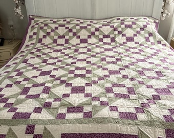 Puede incluir: Un edredón hecho a mano con un patrón geométrico en tonos morado, verde y blanco. El edredón está extendido sobre una cama, mostrando su intrincado diseño y artesanía. El borde del edredón presenta una tela estampada.