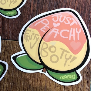May include: Three peach-shaped stickers with the text "Just Peachy Fruity Booty!" in pink and white lettering. The stickers are on a woodgrain surface.