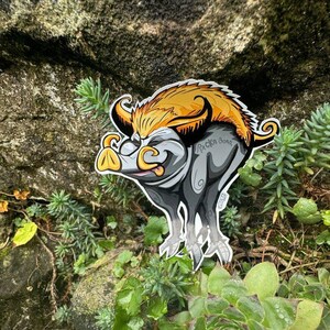 May include: A cartoon sticker of a grey and yellow boar with a curled tail and the text "Rock On?" on its side. The sticker is placed on a rock with green plants growing around it.