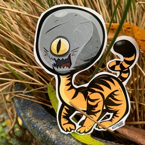 May include: A grey and orange cartoon sticker of a creature with a large, single eye and a tiger-like body. The sticker is on a background of brown grass.