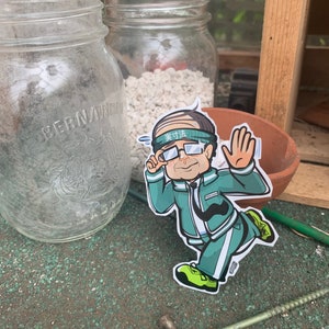 May include: A cartoon sticker of a man wearing a green headband with the text "実力" in white, green glasses, a green jacket, and white pants. He is running with his right arm raised and his left hand holding a fist. The sticker is on a brown surface with a green background.