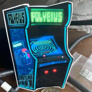May include: A sticker depicting a vintage arcade game called Polybius. The sticker is blue, green and black with a pixelated design. The game is set in Portland, 1981.