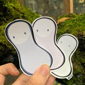 May include: Three ghost-shaped stickers with different coloured outlines. The stickers are white with a swirl design and two black dots for eyes. The stickers are arranged in a stack with the green outlined sticker on the bottom, the purple outlined sticker in the middle, and the blue outlined sticker on top.