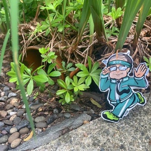 May include: A cartoon sticker of a man wearing a green tracksuit, a headband, and glasses. He is running with his arms outstretched. The sticker is placed on a bed of gravel and green plants.