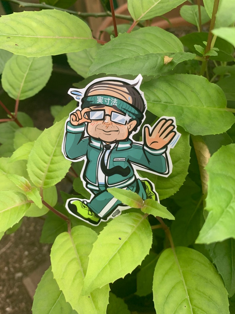 May include: A green and white cartoon sticker of a person wearing a headband with the text "実力派" in Japanese. The person is wearing glasses and a green jacket with a white stripe. The person is holding their hand up in a wave.