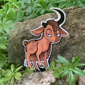 May include: A brown cartoon goat with a single black horn, brown fur, and a stitched scar on its side. The goat has a grumpy expression and is looking to the right. The sticker is on a rock with green plants in the background.