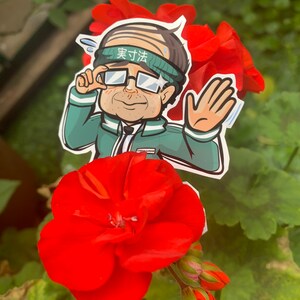 May include: A cartoon sticker of a man wearing glasses and a green headband with the text "実力派" on it. The sticker is placed on a red flower.