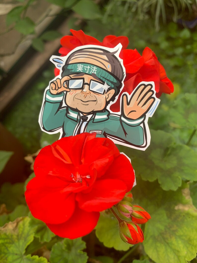 May include: A cartoon sticker of a man wearing glasses and a green headband with the text "実力派" on it. The sticker is placed on a red flower.
