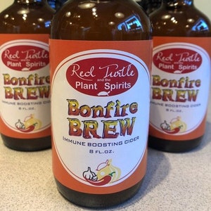 May include: A group of amber glass bottles with labels that say "Red Turtle and the Plant Spirits Bonfire Brew Immune Boosting Cider 8 fl. oz.". The labels have a red, white, and orange color scheme with a graphic of a chili pepper and garlic.