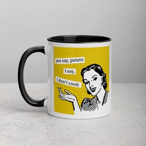Op de afbeelding: Witte keramische mok met een zwart handvat en rand. De mok heeft een retro illustratie van een vrouw en de tekst "you say, potato, I say, I don't cook." De achtergrond is geel met kleine witte stippen.