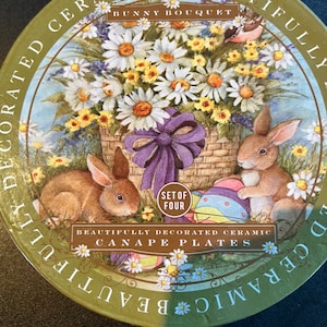 Op de afbeelding: Set van vier prachtig gedecoreerde keramische canapé-bordjes met een konijnenboeket met madeliefjes en een paarse strik. De bordjes zijn groen met gouden letters die "Beautifully Decorated Ceramic Canapé Plates" en "Set of Four" lezen.