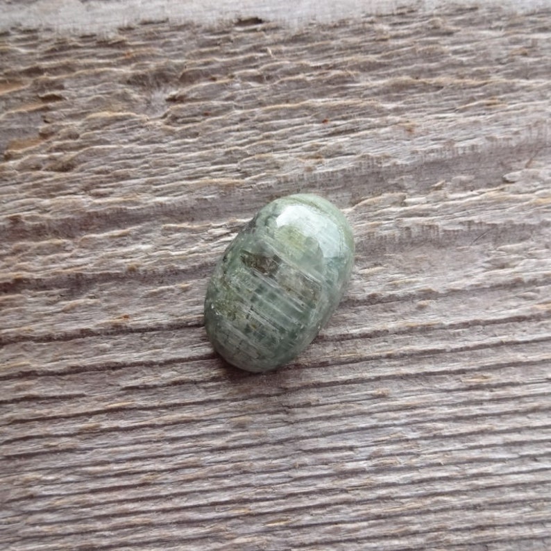 K&ouml;nnte beinhalten: Ein ovaler Cabochon-Edelstein in Gr&uuml;n und Grau mit einer glatten, polierten Oberfl&auml;che. Der Stein hat ein gestreiftes Muster und liegt auf einem h&ouml;lzernen Hintergrund.