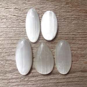 May include: Five polished selenite stones in various shapes: three ovals, one teardrop, and one elongated oval. The stones are white and translucent, with visible internal striations. They rest on a textured, light brown wooden surface.