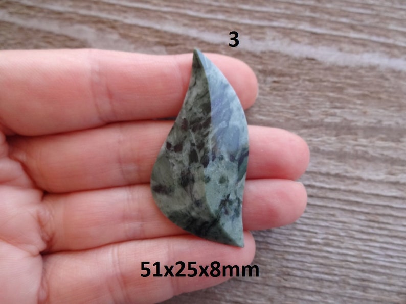 Puede incluir: Un colgante de piedra en forma de hoja, de color verde y negro, que mide 51x25x8mm. La piedra tiene un patr&oacute;n moteado y est&aacute; pulida.