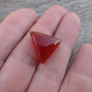 Puede incluir: Una gema triangular pulida, de un rico tono rojo-anaranjado. La gema se sostiene en una mano, mostrando su superficie lisa y reflectante y su forma geom&eacute;trica. El color de la piedra es uniforme.