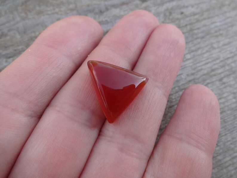 Puede incluir: Una gema triangular pulida, de un rico tono rojo-anaranjado. La gema se sostiene en una mano, mostrando su superficie lisa y reflectante y su forma geom&eacute;trica. El color de la piedra es uniforme.