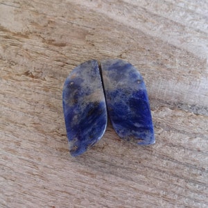 K&ouml;nnte beinhalten: Zwei blaue und wei&szlig;e Lapislazuli-Cabochons, jeder mit einem einzigartigen Muster aus Adern und Wirbeln. Die Steine sind glatt und poliert und haben einen leicht schillernden Glanz.