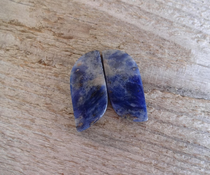K&ouml;nnte beinhalten: Zwei blaue und wei&szlig;e Lapislazuli-Cabochons, jeder mit einem einzigartigen Muster aus Adern und Wirbeln. Die Steine sind glatt und poliert und haben einen leicht schillernden Glanz.