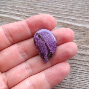 May include: A purple and black gemstone cabochon with a teardrop shape. The stone has a rough, textured surface.