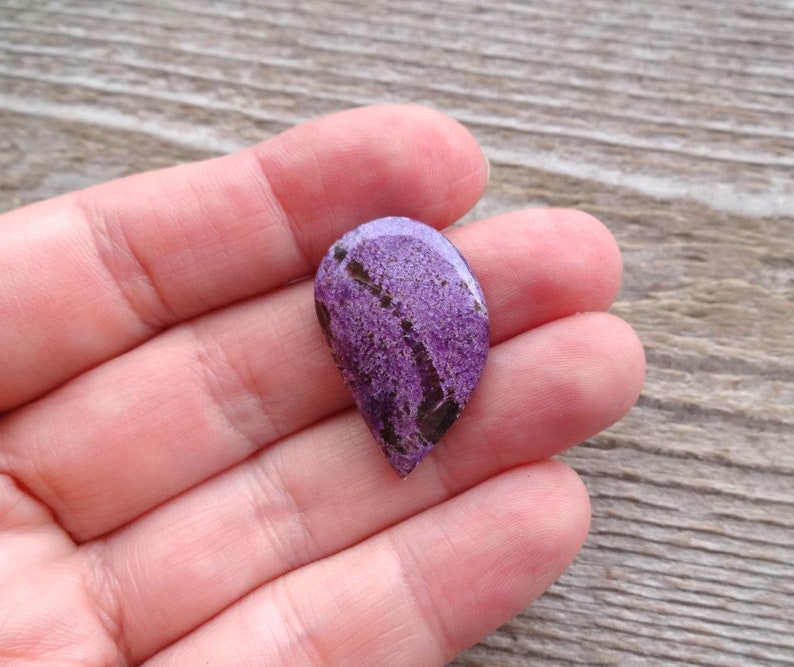May include: A purple and black gemstone cabochon with a teardrop shape. The stone has a rough, textured surface.