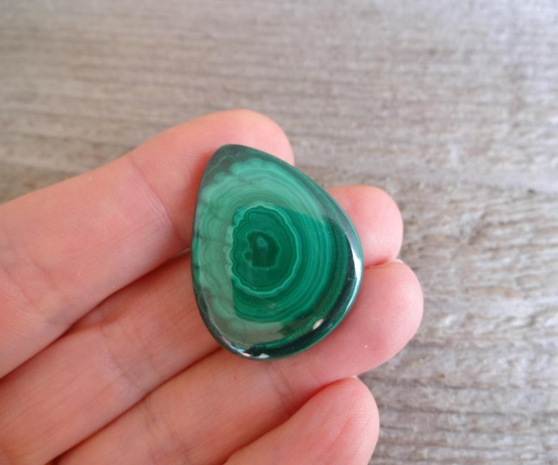K&ouml;nnte beinhalten: Ein gr&uuml;ner Malachit-Cabochon mit einem wirbelnden Muster. Der Cabochon ist tropfenf&ouml;rmig und hat eine glatte, polierte Oberfl&auml;che.