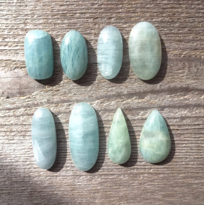 K&ouml;nnte beinhalten: Acht Aquamarin-Cabochons in verschiedenen Formen und Gr&ouml;&szlig;en, darunter oval, tropfenf&ouml;rmig und birnenf&ouml;rmig. Die Steine sind hellblaugr&uuml;n und haben eine glatte, polierte Oberfl&auml;che.
