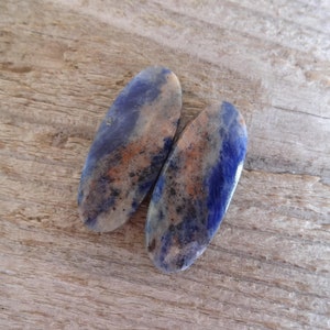 K&ouml;nnte beinhalten: Zwei ovale, blaue, wei&szlig;e und braune Edelsteinkabochons. Die Cabochons haben ein geflecktes Muster und sind glatt und poliert.