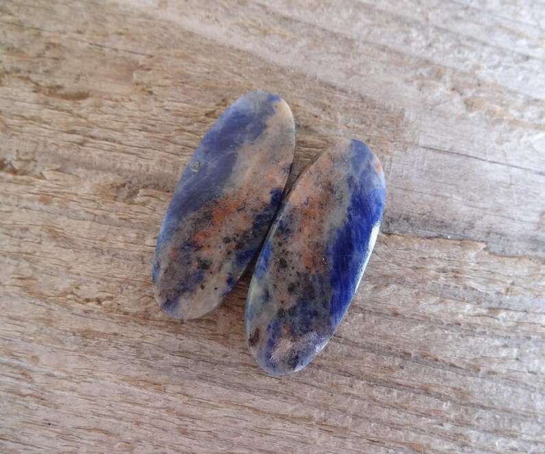 K&ouml;nnte beinhalten: Zwei ovale, blaue, wei&szlig;e und braune Edelsteinkabochons. Die Cabochons haben ein geflecktes Muster und sind glatt und poliert.