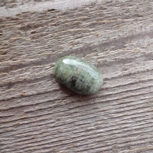 K&ouml;nnte beinhalten: Ein ovaler Cabochon-Edelstein in Gr&uuml;n und Grau mit einer glatten, polierten Oberfl&auml;che. Der Stein hat ein gestreiftes Muster und liegt auf einem h&ouml;lzernen Hintergrund.