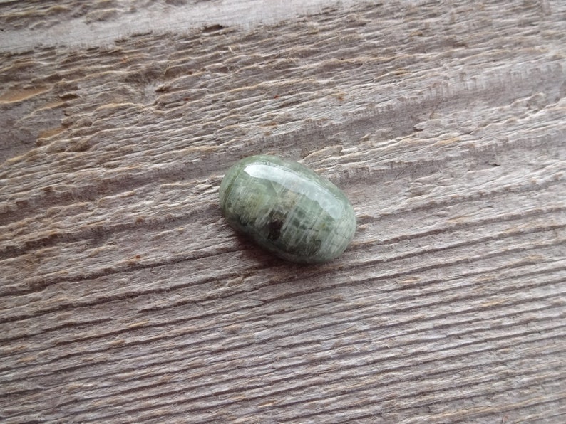 K&ouml;nnte beinhalten: Ein ovaler Cabochon-Edelstein in Gr&uuml;n und Grau mit einer glatten, polierten Oberfl&auml;che. Der Stein hat ein gestreiftes Muster und liegt auf einem h&ouml;lzernen Hintergrund.