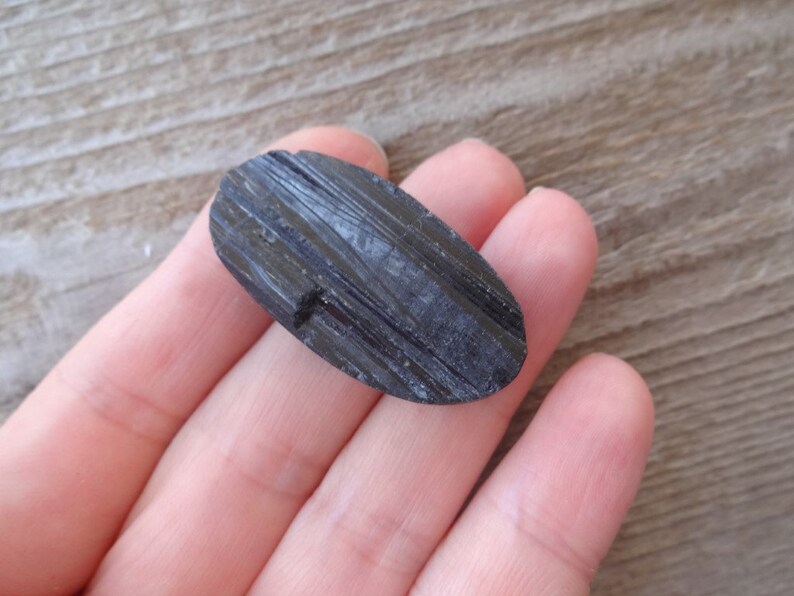 May include: A black, oval-shaped piece of black tourmaline, a type of gemstone, is held in a hand.