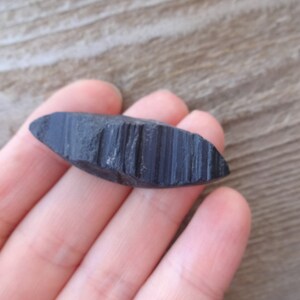 May include: A black, smooth, and slightly curved stone with a striated pattern. The stone is about 3 inches long and 1 inch wide.