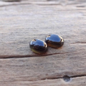 May include: Two oval-shaped, dark brown glass cabochons with a slight amber hue. The cabochons are sitting on a wooden surface.