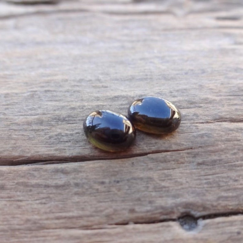 May include: Two oval-shaped, dark brown glass cabochons with a slight amber hue. The cabochons are sitting on a wooden surface.