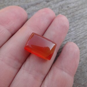Puede incluir: Una gema rectangular pulida con un degradado de tonos naranja y rojo. La piedra se sostiene en una mano, mostrando su superficie lisa y reflectante. La forma de la gema es un rect&aacute;ngulo con esquinas ligeramente redondeadas.