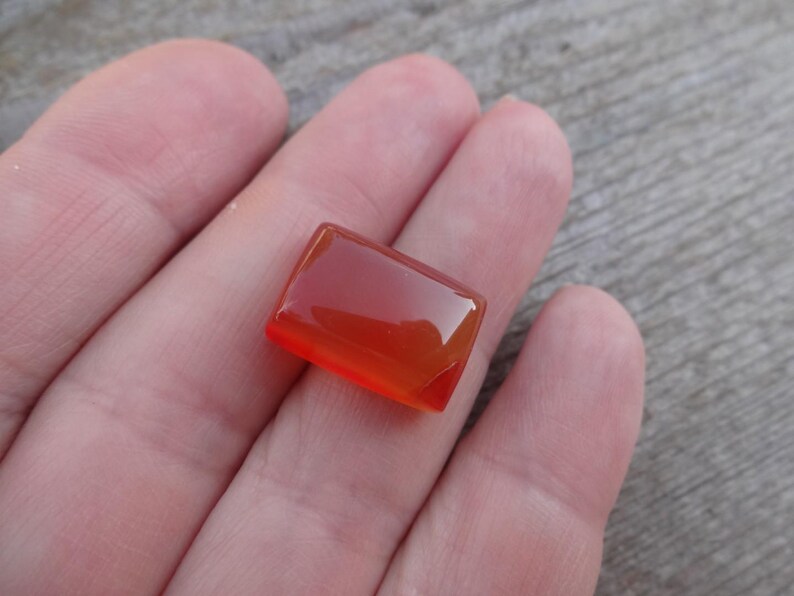Puede incluir: Una gema rectangular pulida con un degradado de tonos naranja y rojo. La piedra se sostiene en una mano, mostrando su superficie lisa y reflectante. La forma de la gema es un rect&aacute;ngulo con esquinas ligeramente redondeadas.