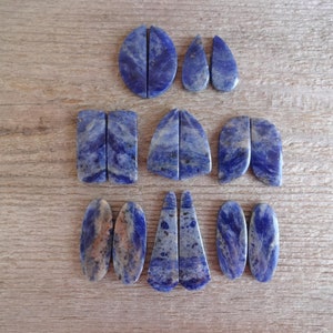 K&ouml;nnte beinhalten: Ein Set aus zehn blauen und wei&szlig;en Edelstein-Cabochons, geschnitten in verschiedenen Formen, darunter oval, tropfenf&ouml;rmig und rechteckig. Die Steine sind in drei Reihen auf einer Holzoberfl&auml;che angeordnet.