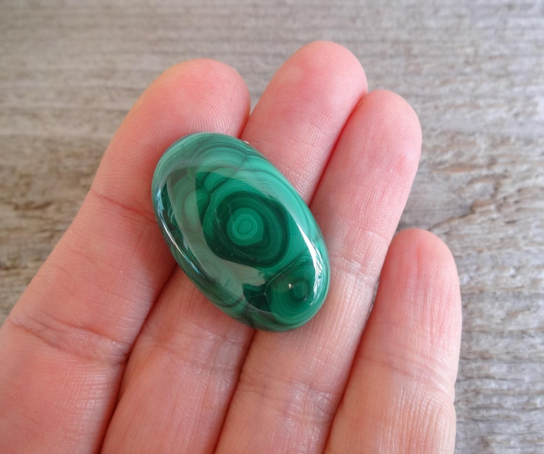 K&ouml;nnte beinhalten: Ein gr&uuml;ner Malachit-Cabochon mit einem wirbelnden Muster. Der Stein ist oval geformt und hat eine glatte, polierte Oberfl&auml;che.